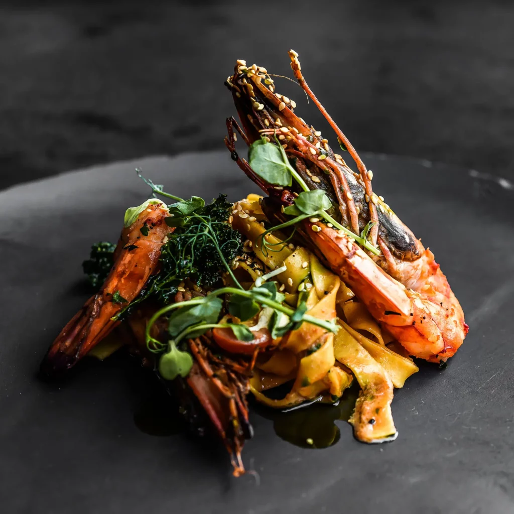 Gambas asadas con pasta y verduras salteadas sobre un plato negro